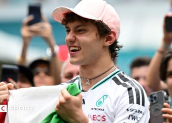 Kimi Antonelli celebrates his race win in Miami while holding the Italian flag. A crowd are gathered around him and taking photographs