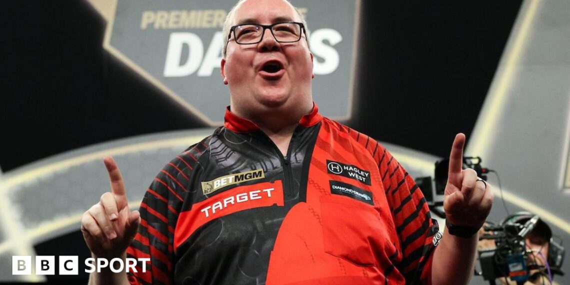 Stephen Bunting celebrates, pointing with both index fingers and pulling a celebratory facial pose as he wears a black and red darts shirt