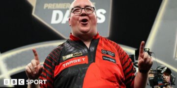 Stephen Bunting celebrates, pointing with both index fingers and pulling a celebratory facial pose as he wears a black and red darts shirt