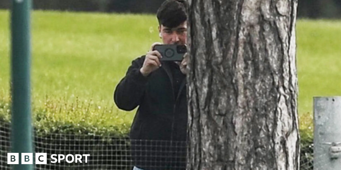 A man in a dark jacket and blue jeans stands partially concealed by a tree trunk and holds a phone up in two hands as if filming and has a white earphone in his right ear