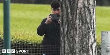 A man in a dark jacket and blue jeans stands partially concealed by a tree trunk and holds a phone up in two hands as if filming and has a white earphone in his right ear