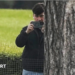 A man in a dark jacket and blue jeans stands partially concealed by a tree trunk and holds a phone up in two hands as if filming and has a white earphone in his right ear