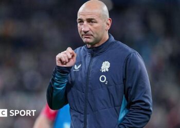 Steve Borthwick, wearing a nany blue zip-up England jacket, looks into the distance, his right hand raised to shoulder level