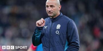 Steve Borthwick, wearing a nany blue zip-up England jacket, looks into the distance, his right hand raised to shoulder level