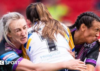 Leeds player Ruby Enright, with a white shirt bearing the number one, is tackled by St Helens players Jodie Cunningham (left) and Zoe Harris (right), both wearing black with purple trim,