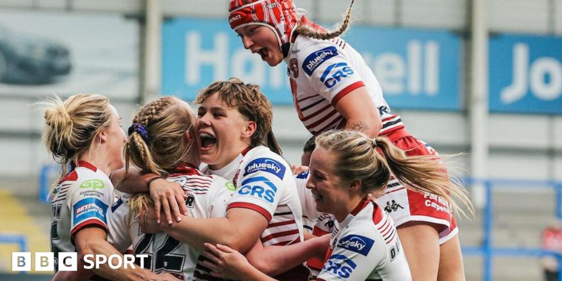 Wigan Warriors women players, in white kit with red trim,  surround try-scorer Ruby Hunter, with one jumping on the shoulders of a team-mate