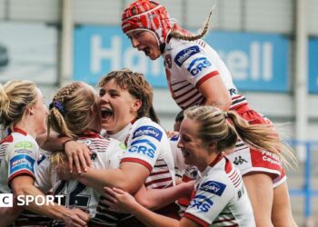 Wigan Warriors women players, in white kit with red trim,  surround try-scorer Ruby Hunter, with one jumping on the shoulders of a team-mate