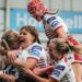 Wigan Warriors women players, in white kit with red trim,  surround try-scorer Ruby Hunter, with one jumping on the shoulders of a team-mate