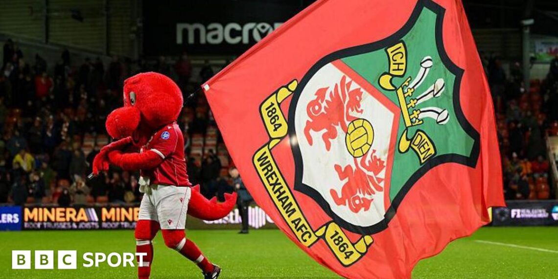 Mascot Wrex the Dragon carries a Wrexham flag across the pitch