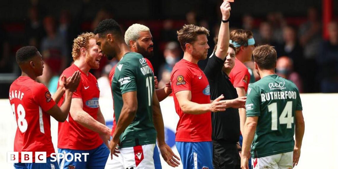 Paul Rutherford is shown a red card with his back to the camera showing his name and number in Wrexham's green away shirt, with Dagenham players in red shirts around the referee.