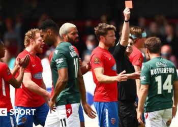 Paul Rutherford is shown a red card with his back to the camera showing his name and number in Wrexham's green away shirt, with Dagenham players in red shirts around the referee.