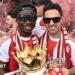Bukayo Saka and Mikel Arteta, both in sunglasses and red and white team kit, with the Premier League trophy on Arsenal's bus parade