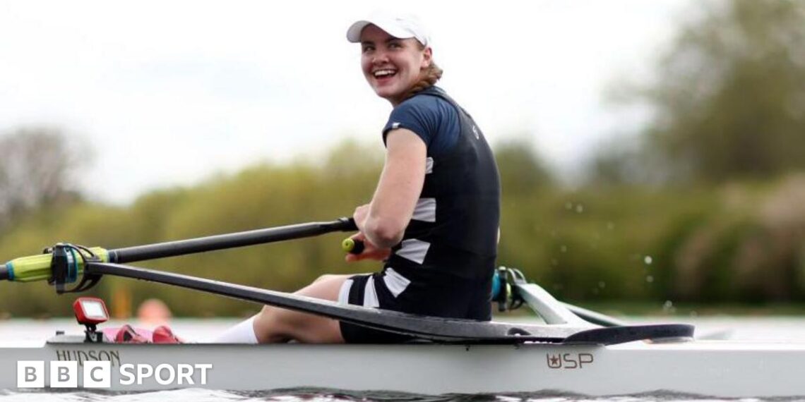 Lauren Henry turns and smiles while in her boat on the water, wearing a dark rowing outfit and white cap