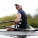 Lauren Henry turns and smiles while in her boat on the water, wearing a dark rowing outfit and white cap