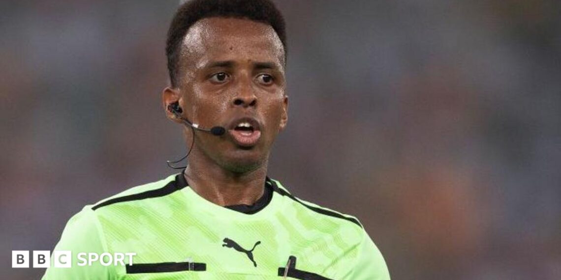 Omar Artan refereeing a match, with earpiece and microphone, and a Fifa badge on his light green shirt