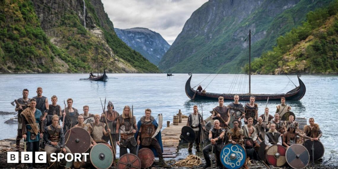Norway players in Viking dress and with weapons and shields pose in front of a fjord with Viking boats