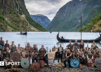 Norway players in Viking dress and with weapons and shields pose in front of a fjord with Viking boats