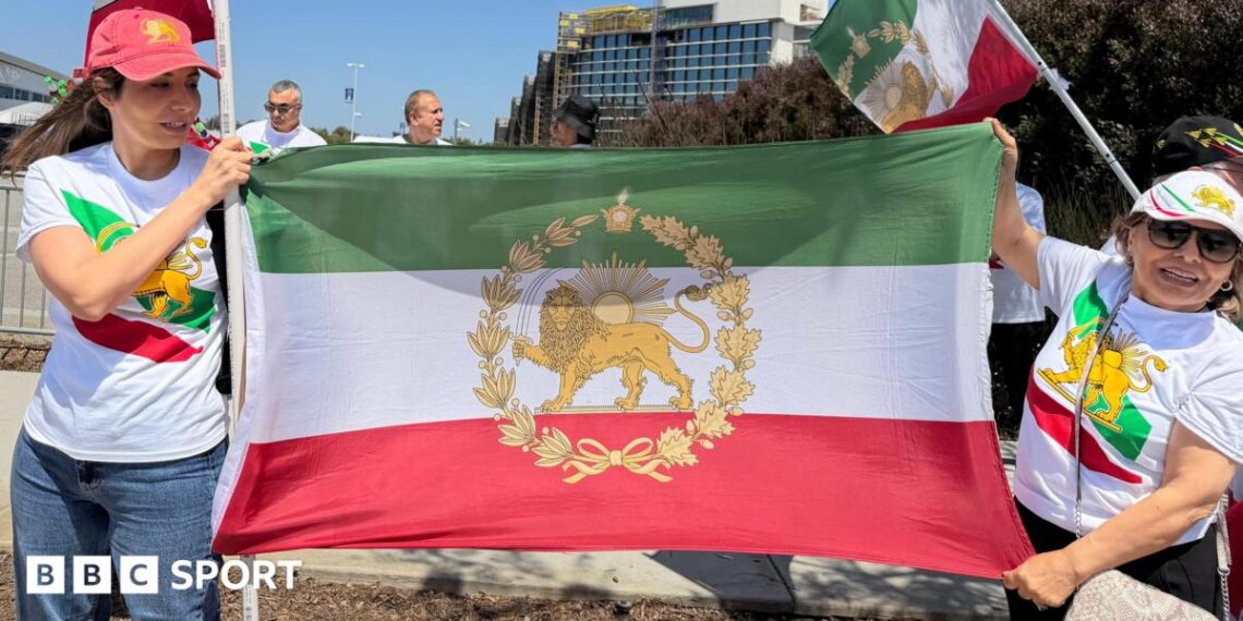 Two women wear t shirts and caps featuring the stylised lion symbol of the flag of Iran's pre-1979 Islamic revolution. They are also holding the flag between them.