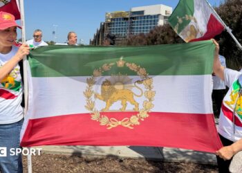 Two women wear t shirts and caps featuring the stylised lion symbol of the flag of Iran's pre-1979 Islamic revolution. They are also holding the flag between them.